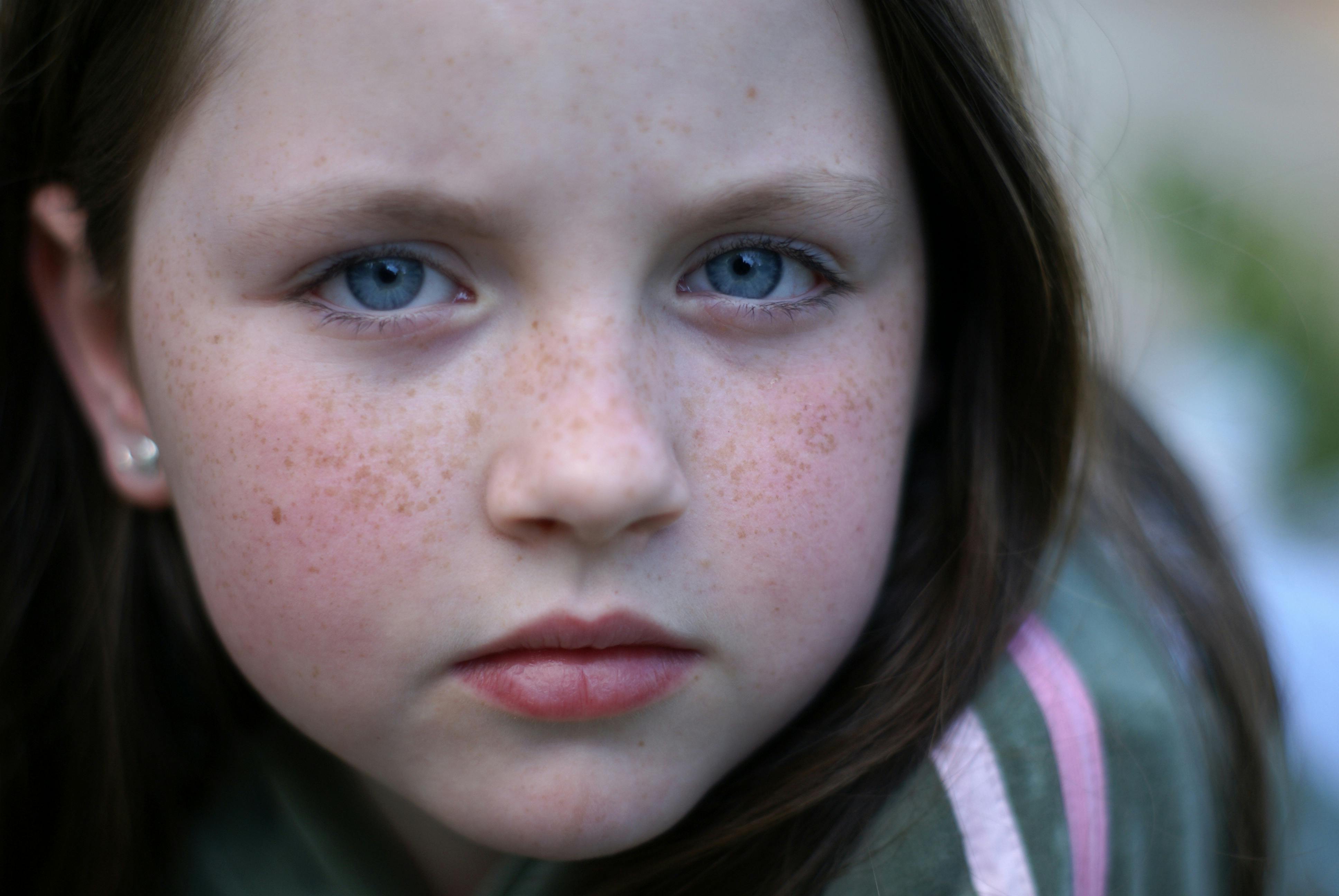 A detailed close-up portrait of a young girl with freckles and blue eyes, conveying a serious expression.